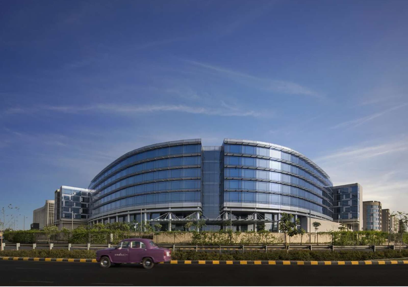 The curved glass facade of Andaz Delhi in Aerocity under a clear blue sky, with a vintage Ambassador taxi passing on the road in front.