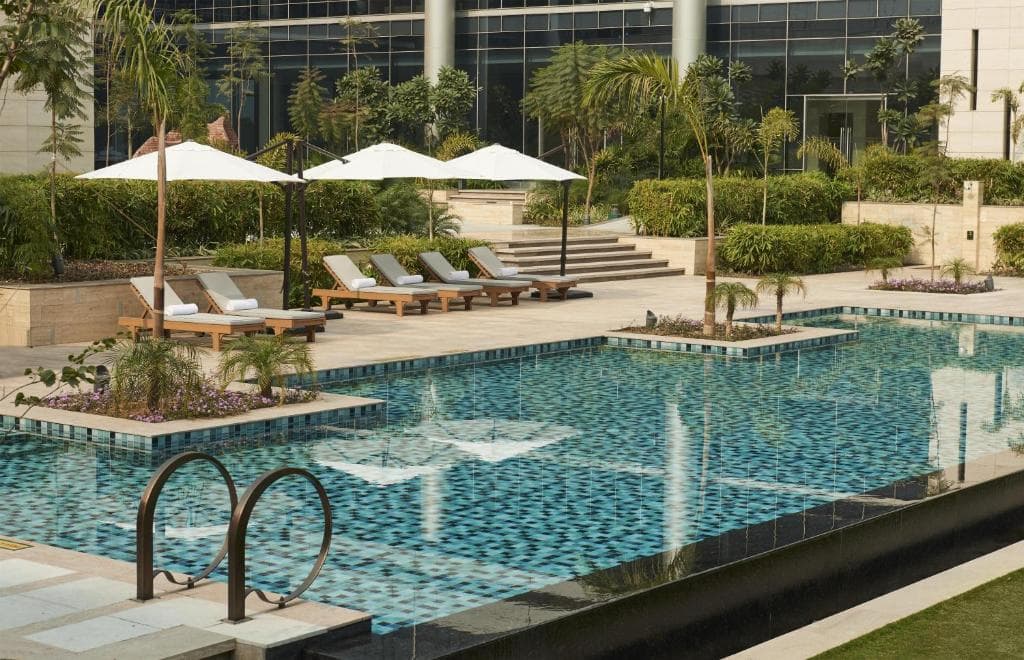 The outdoor pool at Andaz Delhi lined with wooden loungers and white umbrellas, the hotel's curved glass wing visible behind.
