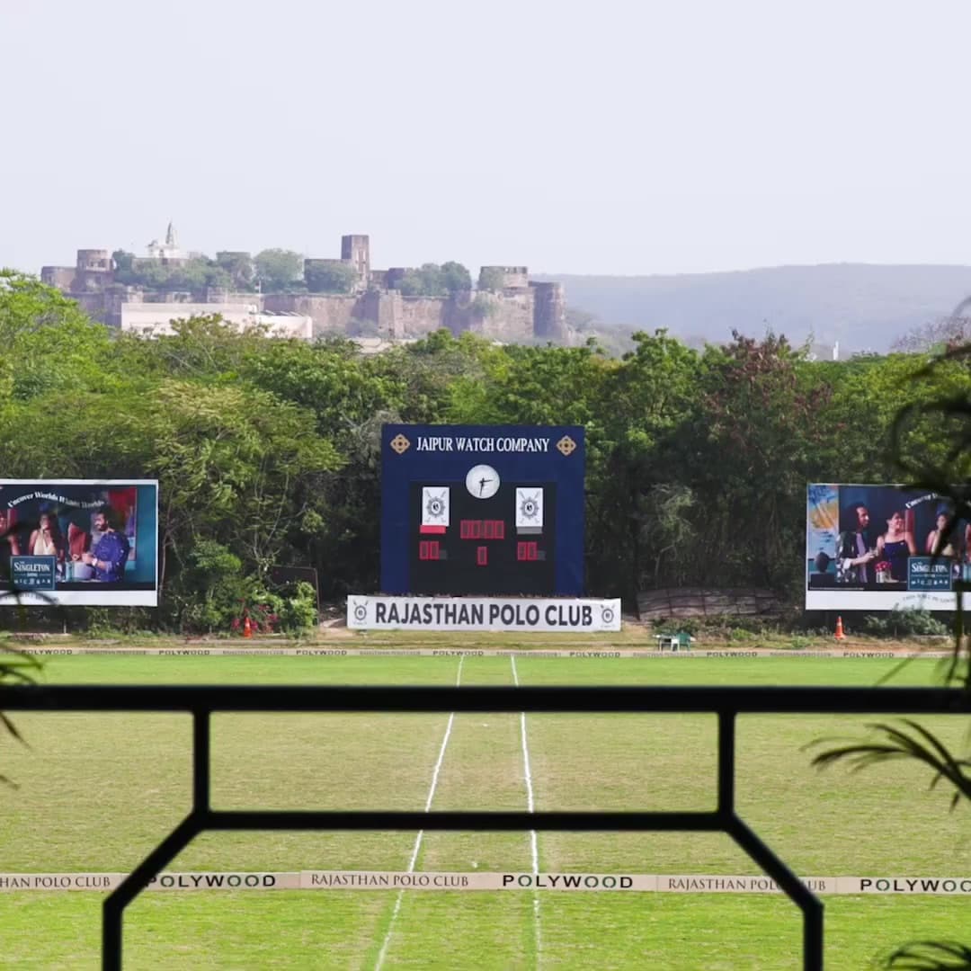 The Rajasthan Polo Club pitch in Jaipur, goalposts and an LED scoreboard reading Rajasthan Polo Club, with a hilltop fort visible beyond the treeline.